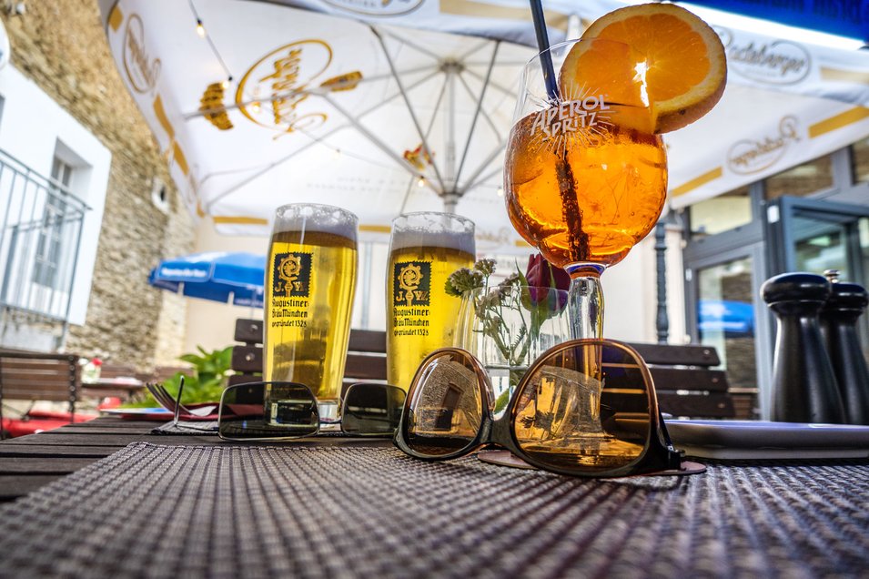 Ein Tisch im Biergarten mit einem Spritz-Getränk und Bieren, davor liegen Sonnenbrillen. Die Sonnenschirme schützen vor der Sonne.